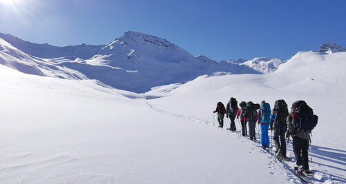 Eine Gruppe Kollegiaten wandert hintereinander durch eine weiße, bergige Schneelandschaft 