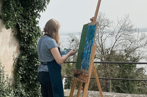 Eine Kollegiatin steht mit Aussicht auf dem Bodensee vor eine Leinwand und malt ein Gemälde.