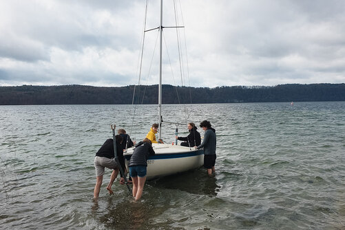 Sechs Kollegiatinnen und Kollegiaten beim einwässern eines Segelboots. Im Hintergrund ist eine Hügellandschaft über dem Bodensee zu erkennen.  