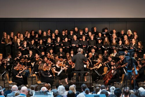Orchesteraufführung: Thomas Braun dirigiert, Streicher spielen, Chor singt.