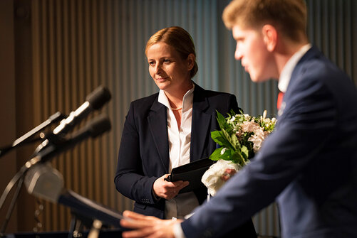 Claudia Groot und Kollegiat mit Blumenstrauß am Rednerpult bei Abschlussfeier.