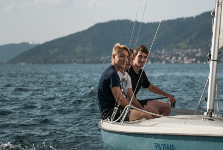 Zwei Jugendliche sitzen auf einem Segelboot auf dem Bodensee bei schönem Wetter. 
