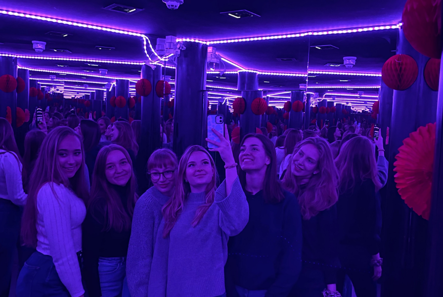 Zu sehen sind sechs Kollegiatinnen die in einem Spiegel ein Gruppenfoto machen. Der Hintergrund ist lila beleuchtet.