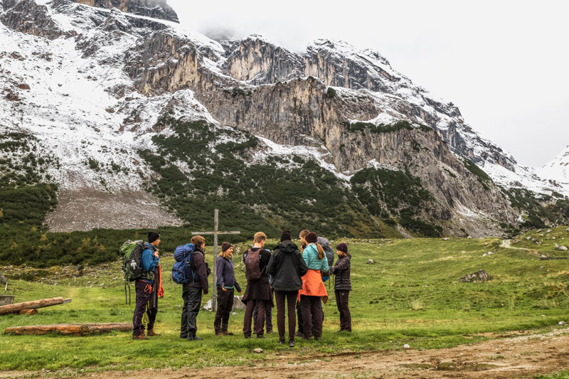 Eine Gruppe steht im Kreis in einem Tal umringt von leicht schneebedeckten Bergen