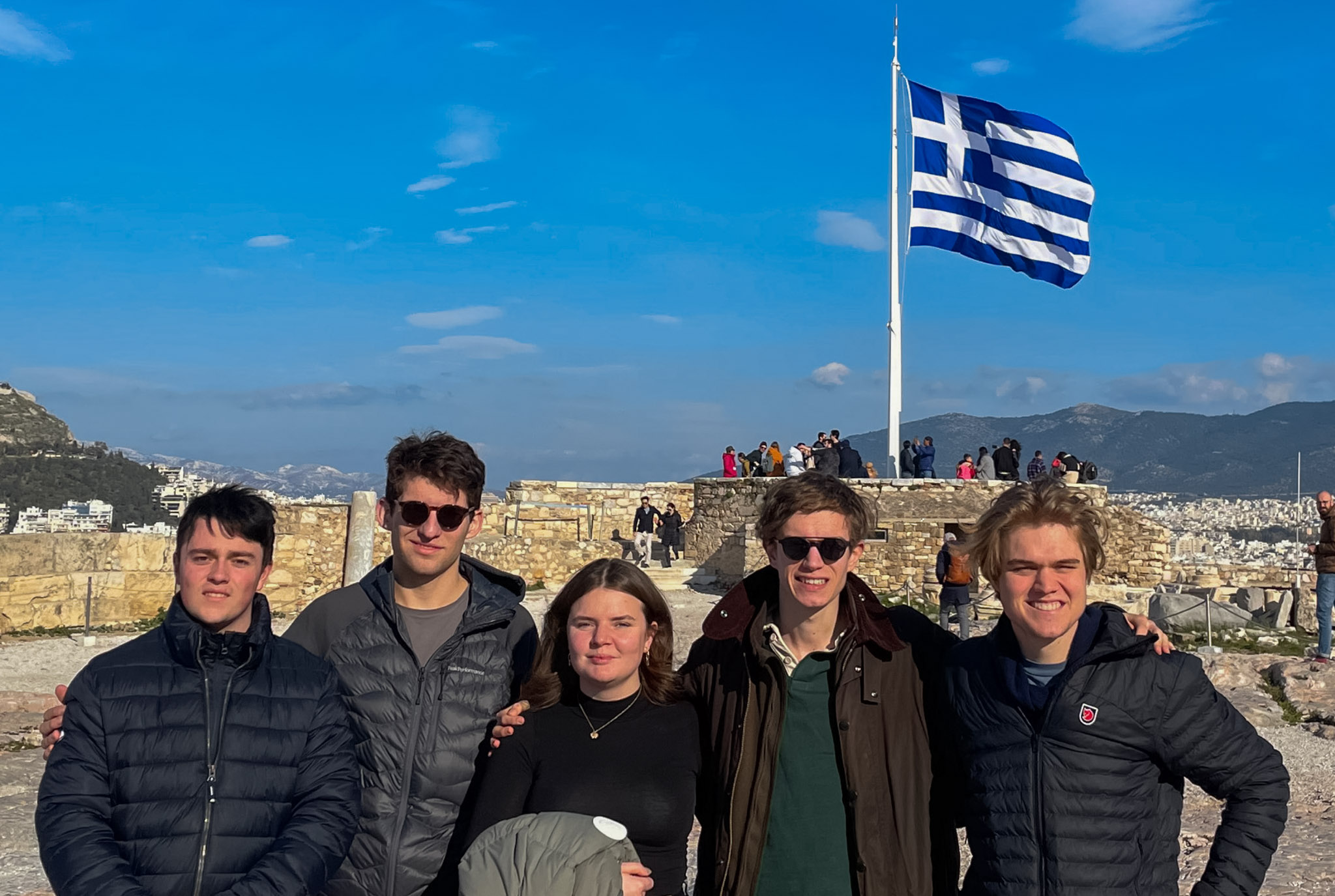 Projektgruppe steht vor einer griechischen Fahne hinter der Akropolis in Athen.