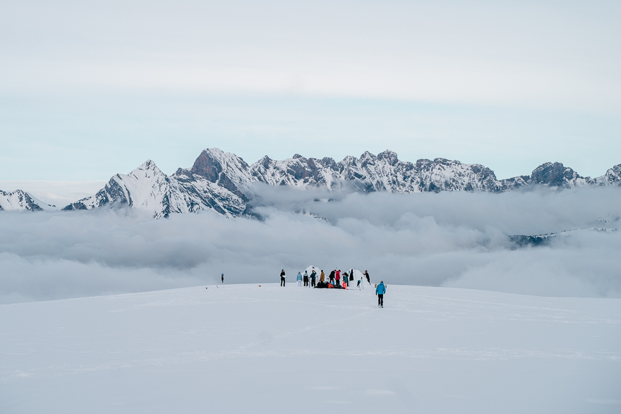 Alpen tauchen aus den Wolken auf, Kollegiaten bauen Iglus im Vordergrund.