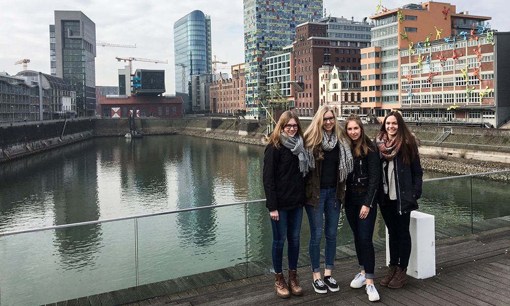 Vier Kollegiatinnen stehen auf einer Brücke mit dem Roggendorfer Haus in Düsseldorf im Hintergrund.