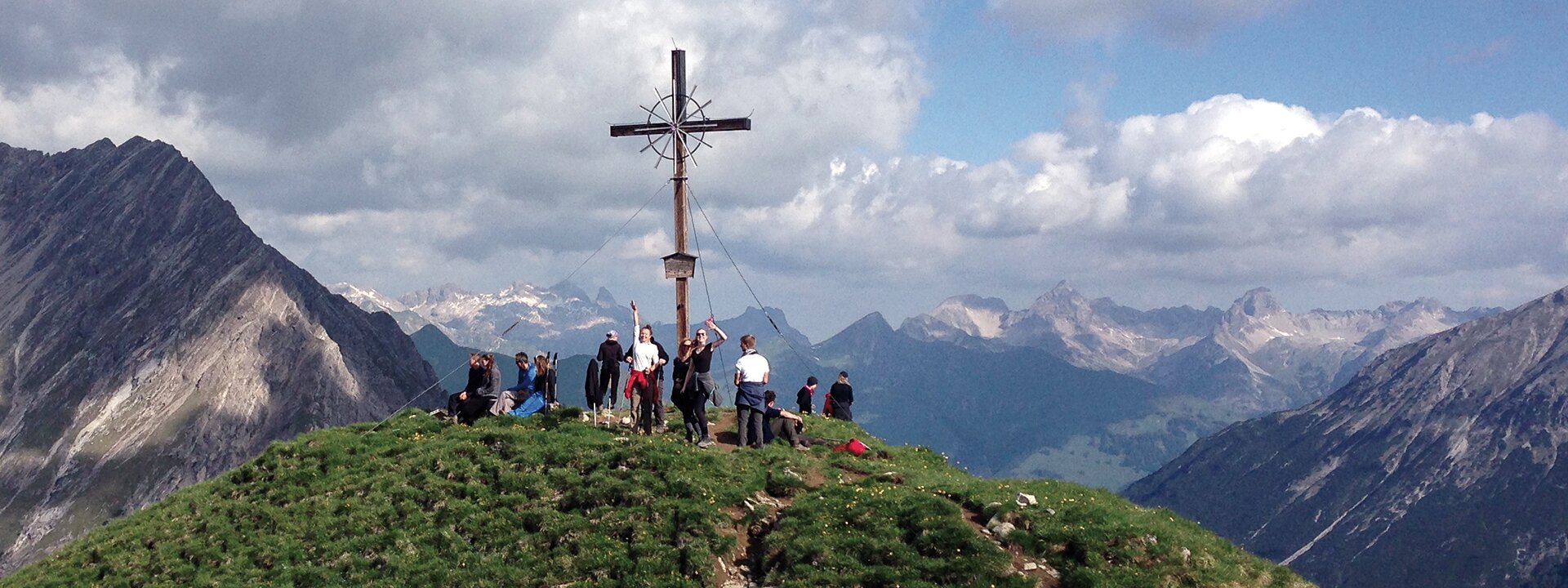 Weite Aufnahme einer Gruppe während einer Exkursion unter dem Gipfelkreuz vor einem Bergpanorama